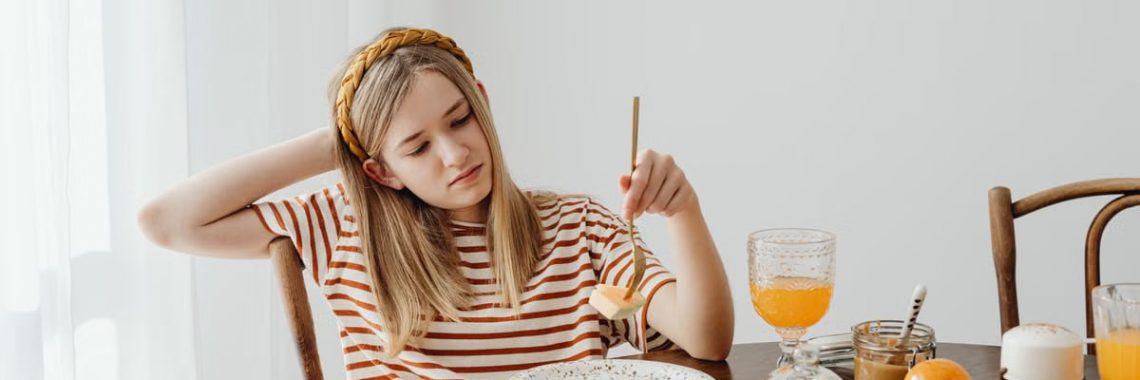 Teen girl in striped shirt at breakfast table with fruits, bread, and juice. Casual morning setting.