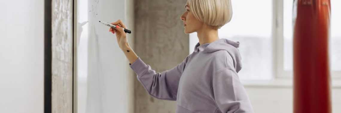 Fit female trainer writes workout plan on a whiteboard in a gym setting. Side view.